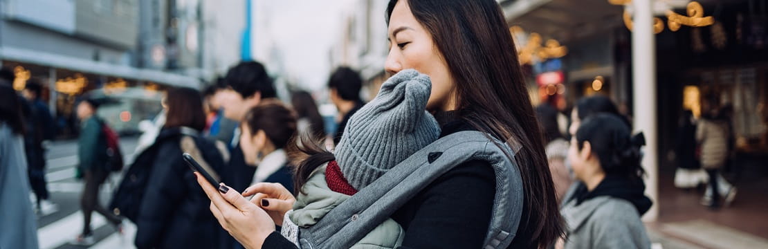 A woman walking in a metropolitan street, looking at her phone. She is holding a baby in a front carrier.